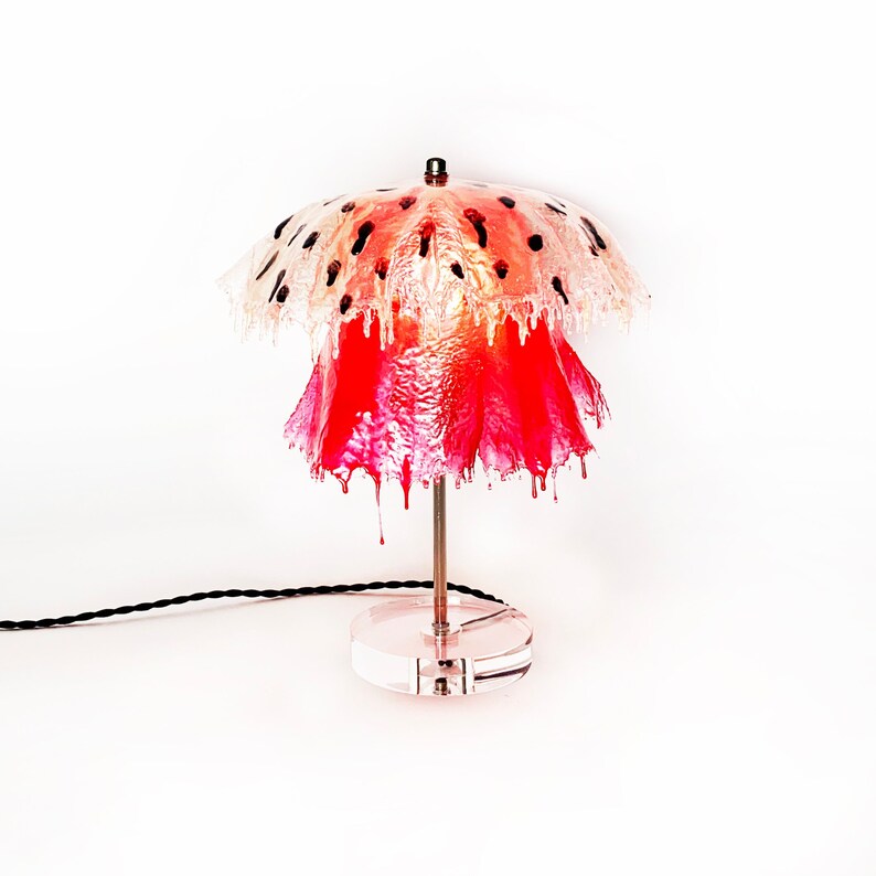 May include: A table lamp with a pink and white lampshade that resembles a jellyfish. The lampshade has a clear, translucent top with black spots and a pink, opaque bottom. The lamp has a clear acrylic base.