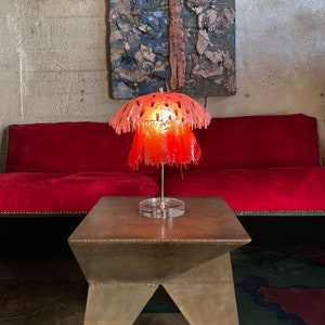 May include: A red, dripping, lampshade with a geometric pattern sits on a brown, metal table. The lamp is lit and casts a warm glow.