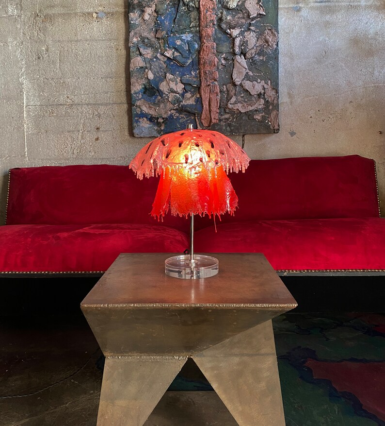 May include: A red, dripping, lampshade with a geometric pattern sits on a brown, metal table. The lamp is lit and casts a warm glow.
