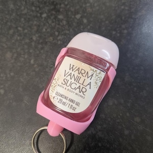 May include: A pink hand sanitizer bottle with a white cap and a pink holder. The label reads "Warm Vanilla Sugar Cleansing Hand Gel" and contains 29 ml / 1 fl oz. A silver key ring is attached to the holder.