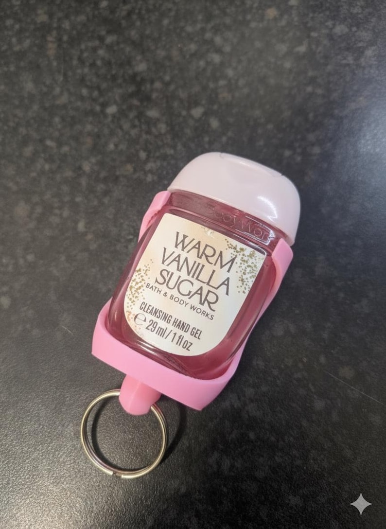 May include: A pink hand sanitizer bottle with a white cap and a pink holder. The label reads "Warm Vanilla Sugar Cleansing Hand Gel" and contains 29 ml / 1 fl oz. A silver key ring is attached to the holder.