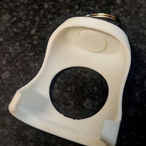 May include: A white, plastic key fob holder with a circular opening and a small, oval-shaped pad. A metal keyring is attached to the top. The holder has a curved design and is likely used to protect or organize keys.
