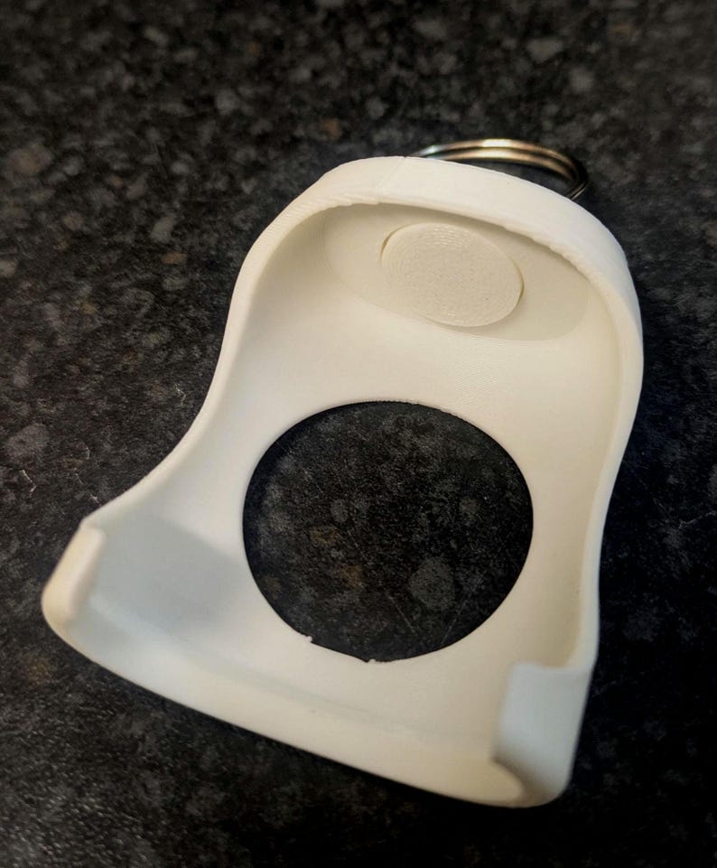 May include: A white, plastic key fob holder with a circular opening and a small, oval-shaped pad. A metal keyring is attached to the top. The holder has a curved design and is likely used to protect or organize keys.