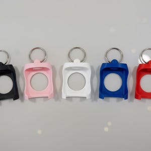May include: Five colorful keychains in black, pink, white, blue, and red. Each keychain has a circular opening and a silver metal ring at the top for attaching keys. The keychains are made of a smooth, solid material.