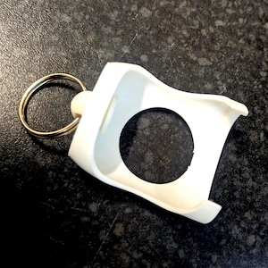 May include: A white, plastic keychain holder with a circular opening and a gold-colored key ring. The holder has a curved design and is likely used to secure a small item, such as a smart tag or a small electronic device.