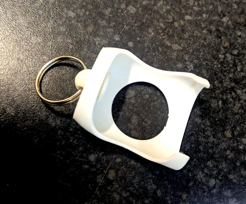 May include: A white, plastic keychain holder with a circular opening and a gold-colored key ring. The holder has a curved design and is likely used to secure a small item, such as a smart tag or a small electronic device.