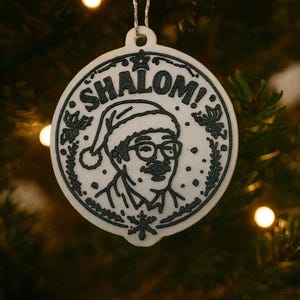 Könnte beinhalten: Ein weißer, kreisförmiger Weihnachtsschmuck mit schwarzer Umrandung. Er zeigt ein Porträt eines Mannes mit Weihnachtsmütze und Brille, darüber das Wort "SHALOM!". Der Schmuck hängt an einem silbernen Faden.