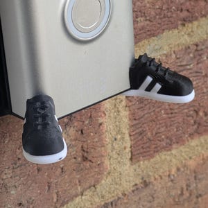 Puede incluir: Un timbre plateado con un botón circular, adornado con dos zapatillas deportivas en miniatura en blanco y negro. Las zapatillas tienen rayas y suelas blancas, montadas en una pared de ladrillo rojo.