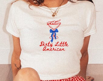 May include: White t-shirt with a cocktail graphic in red and blue, and the text "Dirty Little American." The shirt is paired with red and white checkered shorts and red cowboy boots with white star accents.