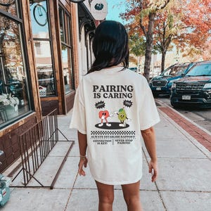 May include: A white t-shirt with a graphic of a red apple and a green pear holding hands. The text on the shirt reads "Pairing is Caring" and "Always Establish Rapport! Connection is Key! Never Stop Pairing!"