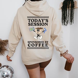 Op de afbeelding: Een beige hoodie met een afbeelding van een koffiekopje dat een rekenmachine en een rietje vasthoudt. De tekst op de hoodie luidt "Today's Session Sponsored by Coffee".