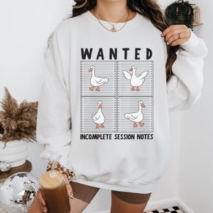 Puede incluir: Sudadera blanca con un gráfico de cuatro gansos de dibujos animados en una alineación policial, con la palabra "WANTED" arriba e "INCOMPLETE SESSION NOTES" abajo. Los gansos tienen patas y picos naranjas.