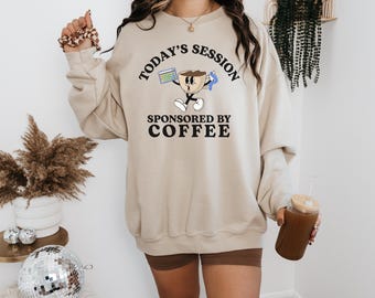 May include: Beige sweatshirt with the text "Today's Session Sponsored by Coffee" and a cartoon coffee cup character. The sweatshirt is paired with brown shorts. A glass of iced coffee is held in the hand.