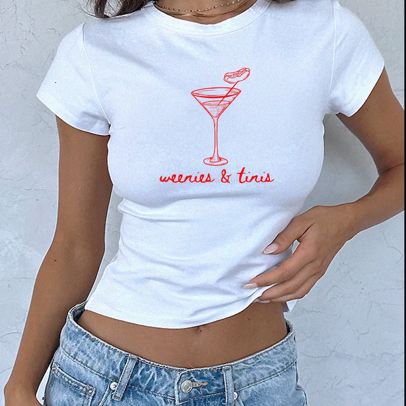 May include: White t-shirt with a red graphic of a martini glass and a hot dog garnish. The text "weenies & tinis" is written in red script below the graphic. The shirt has short sleeves and a crew neck.