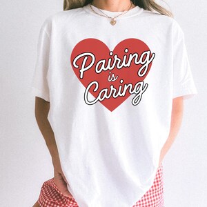 Może przedstawiać: Biała koszulka z czerwonym sercem z napisem "Pairing is Caring" w kursywie. Tekst jest obramowany na czarno. Swobodny top, odpowiedni do noszenia na co dzień.