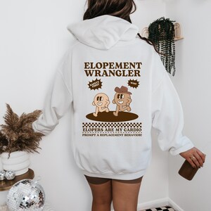 Op de afbeelding: Witte hoodie met de tekst "ELOPEMENT WRANGLER" in bruin. Onder de tekst staat een cartoonafbeelding van twee figuren en de zin "ELOPERS ARE MY CARDIO PROMPT A REPLACEMENT BEHAVIOR!"