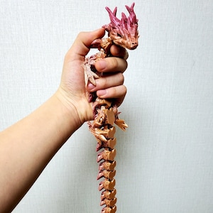 May include: A detailed, articulated dragon figurine in shades of brown, gold, and pink. The dragon is held in a hand, showcasing its segmented body and intricate design. The dragon's head features horns and a detailed face.