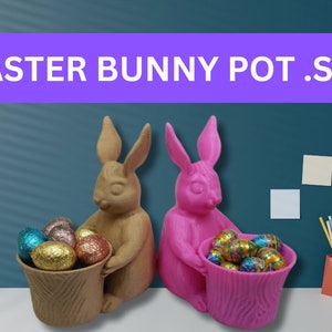 Peut inclure: Deux pots en forme de lapin de Pâques, l'un brun et l'autre rose, contenant des œufs en chocolat. Les lapins sont assis sur une table avec un fond bleu.