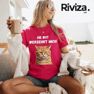 May include: A magenta t-shirt with a grumpy orange tabby cat graphic and the German text "DIE WUT VERZEHRT MICH" in white. The shirt is worn by a person.