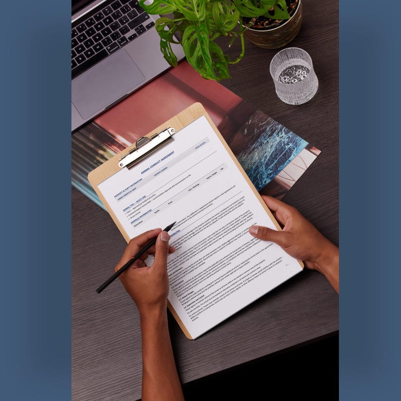 May include: A light-colored wooden clipboard holding a white document titled Animal Conduct Agreement, featuring printed text and a metal clip at the top. A black pen is positioned on the paper, which contains structured sections for information.