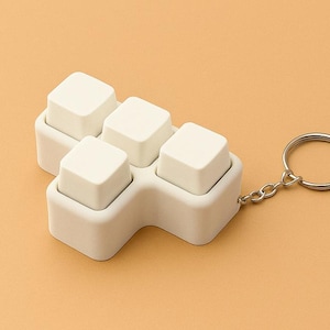 May include: A white, five-button keyboard keycap keyring. The keycaps are arranged in a plus shape and attached to a silver keyring. The background is a solid, light orange colour.