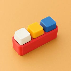 May include: A red rectangular tray holds three square blocks: white, yellow, and blue. The blocks are evenly spaced and sit flush within the tray. The background is a solid orange color.