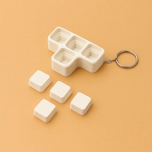 May include: A white, geometric-shaped keychain with a silver ring. The keychain has a series of square compartments, with five detached square pieces. The items are set against a solid, light orange background.