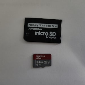 May include: A black Memory Stick PRO Duo compatible micro SD adaptor sits above a red and gray Sandisk Ultra 64GB micro SD card. The cards are on a white surface.