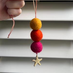 May include: A colorful felt ball garland with a gold starfish charm. The garland features a wooden bead at the top, followed by yellow, orange, and fuchsia felt balls. The starfish charm hangs at the bottom, suspended by a thin string.