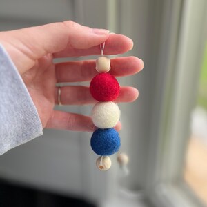 May include: A handmade felt ball garland featuring red, white, and blue balls, strung together with a thin white string. The garland includes small wooden beads. The color scheme is reminiscent of the American flag.