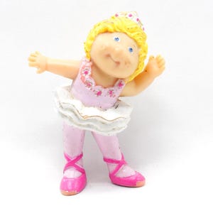 Puede incluir: Pequeña figura de bailarina con pelo rubio y ojos azules. Lleva un maillot rosa, medias y zapatillas de ballet. Un tutú blanco con ribete dorado completa el conjunto. La figura está posando con los brazos extendidos.