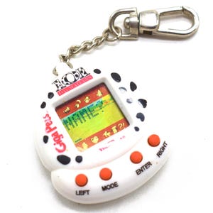 May include: A white Giga Pets digital pet keychain with black spots, featuring a screen displaying the text NAME? and various icons. The device has three orange circular buttons labeled LEFT, MODE, and ENTER, and a metal chain with a lobster clasp.