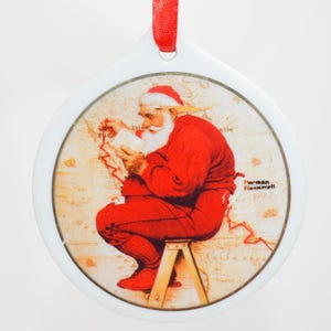 Puede incluir: Adorno navideño de cerámica blanca con una cinta roja. El adorno presenta una ilustración vintage de Papá Noel con un traje rojo, sentado en un pequeño taburete de madera, examinando un mapa. La ilustración está enmarcada por un borde blanco.