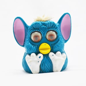 May include: A blue and pink Furby toy with large, round eyes and a yellow beak. The toy has white feet and a white tuft of fur on its head.