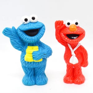 Könnte beinhalten: Zwei Sesame Street-Charakterfiguren. Krümelmonster ist blau und hält einen gelben Buchstaben "E". Elmo ist rot mit einem weißen Lätzchen. Beide haben große Augen und offene Münder, vor weißem Hintergrund.