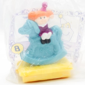 Puede incluir: Figura de juguete vintage de un niño montado en un caballito balancín azul, sobre una base amarilla, dentro de una bolsa de plástico transparente. El niño tiene el pelo naranja y viste un atuendo morado. La bolsa tiene el número 8.