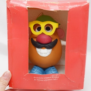 Könnte beinhalten: Ein Mr. Potato Head-Ornament in einer roten Box. Das Spielzeug hat einen braunen Körper, gelbe Brille, eine rote Nase, einen schwarzen Schnurrbart und blaue Schuhe. Auf der Box steht "An exclusive ornament designed by Midwest of Cannon Falls."