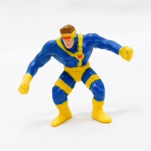 May include: A small, plastic action figure of a superhero in a dynamic pose. The figure is primarily blue with yellow accents, including boots, gloves, and a belt. The character has a brown head and a red visor. The figure is standing on a white surface.