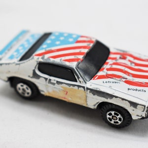 May include: A white toy car with an American flag design. The car has black wheels and a black interior. The car is weathered and has some paint chips. The car has the text "Letraset products" on the side.