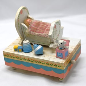 Könnte beinhalten: Eine Vintage-Holzspieluhr mit einem Baby in einer Wiege. Die Box ist in Pastellfarben mit floralen Akzenten bemalt. Enthält einen kleinen weißen Bären, blaue und gelbe Blöcke und andere dekorative Elemente. Ein charmantes Dekorationsstück für das Kinderzimmer.