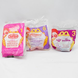 May include: Three sealed McDonald's Happy Meal toys in plastic packaging. The packages are pink, purple, and white, with images of Ronald McDonald and various characters. Each package has a number.