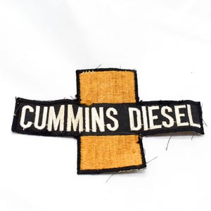 Op de afbeelding: Een rechthoekige geborduurde patch met een horizontale zwarte balk met de witte tekst CUMMINS DIESEL in het midden, doorkruist door een verticaal oranje rechthoekig blok met een zwarte rand, wat een kruisvorm creëert.