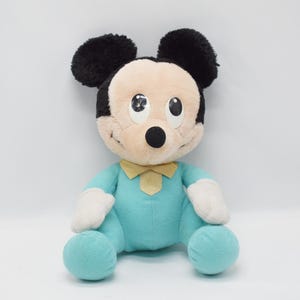 Op de afbeelding: Een pluche Mickey Mouse pop, met zwarte oren, een bruin gezicht en een turquoise outfit met een gele vlinderdas. De pop heeft witte handschoenen en turquoise voeten. Het speelgoed zit op een witte achtergrond.