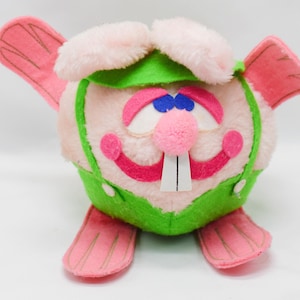 Puede incluir: Un conejito de peluche con orejas, pies y detalles rosas. El conejito tiene una nariz de pompón rosa, ojos azules y un atuendo verde. La boca del conejito es un diseño rojo sonriente con dos dientes blancos.