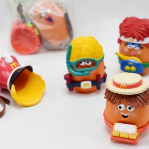 May include: A collection of McDonald's Happy Meal toys featuring anthropomorphic food characters. The toys include a red band uniform hat, a drum, and several figures with various accessories, such as a diver's mask and a straw hat.