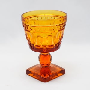 May include: Amber-colored vintage goblet with a square base and a round, patterned bowl. The stem is round with a small ball in the middle. The glass has a textured design around the rim and on the bowl. The goblet is made of glass.