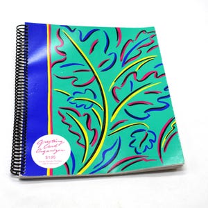 Puede incluir: Un organizador de tarjetas de felicitación con espiral, con una cubierta verde azulado que presenta un diseño abstracto colorido de hojas y líneas en rosa, amarillo y azul. Una franja azul recorre el lomo. Una pegatina blanca con texto está en el frente.