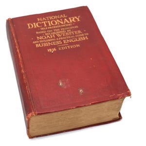 May include: A vintage red dictionary titled "National Dictionary" with gold lettering. The book, published in 1936, includes a guide to business English. The cover shows signs of wear, with visible scuffs and age spots, suggesting its historical use.