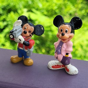 May include: Two small plastic figurines of Mickey Mouse. One Mickey Mouse is wearing a red shirt, blue pants, and a backpack. He is holding a camera. The other Mickey Mouse is wearing a pink jacket, purple pants, and white shoes.