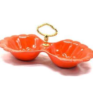 May include: An orange, two-section serving dish with a scalloped edge design. It features a gold-colored handle in the center for easy carrying. The dish is made of a glossy material and is set against a white background.